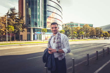 A young man is looking happy while standing outside, on the sidewalk, while behing him there is a big mallの写真素材