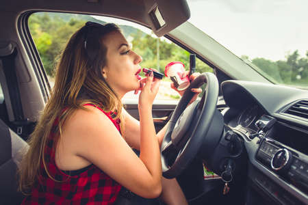Young reckless driving is sitting in her car while is doing her makeup on the car mirror