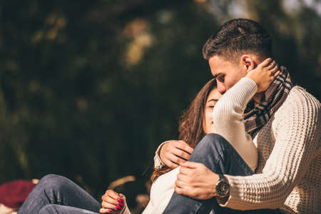 A couple is in the park on a autumn day, the boy is kissing his girlfriend on the head
