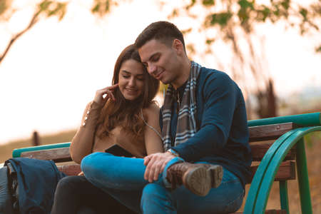 Young couple listening to music,smiling,looking at the phone and sitting on a bench in park.の写真素材