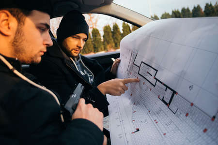 Two robbers planning and pointing on the blueprint(map) their objective while holding their guns.