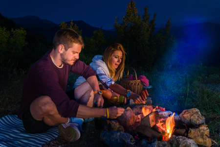 Man and woman rosting sausages on the campfire, drinking beer and relaxing by the campfire in the mountain top in the night. Romantic campfire concept.の写真素材