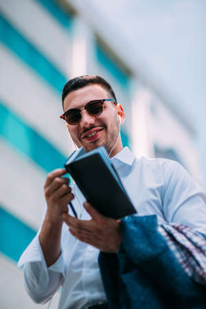 A businessman in suit is looking around him in the city while he holds a pen and opened notebookの写真素材