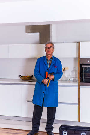 Mature worker standing near toolbox in the kitchen while holding hammer in hs handsの写真素材