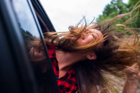 A girl is with her head out of a car window while is driving in nature, smiling and having funの写真素材