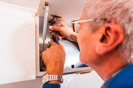 Handyman is in the kitchen and he is fixing the cupboard with screwdriver