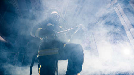 Portrait of a female firefighter while holding an axe and wearing an oxygen mask indoors surrounded...