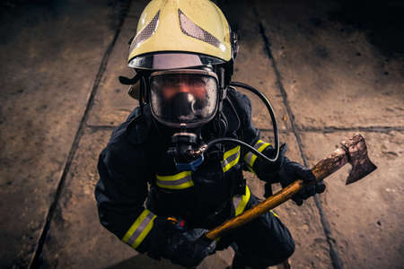 Portrait of a female firefighter while holding an axe and wearing an oxygen mask indoors surrounded...