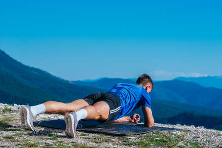 Strong male athlete doing plank while exercising his abs on top of a mountain on a sunny day.