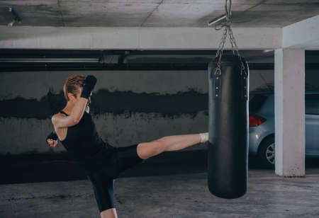 Caucasian woman in sportswear and with black bandages kicking the boxing bag inside a garageの写真素材