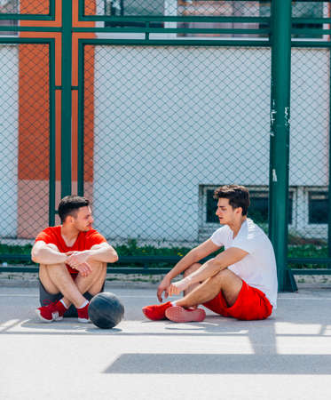 Two strong caucasian athletes resting on the ground at the basketball court while having a conversation.の写真素材