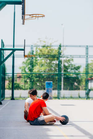 Two strong caucasian athletes resting on the ground at the basketball court while having a conversation.の写真素材