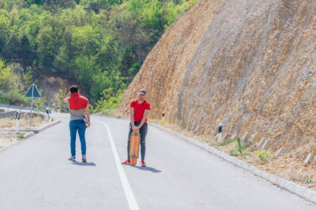 Two male longboarders carrying their longboards in their hands while climbing uphill and preparing for a downhill slide. Wearing red t-shirts, green hat, and super cool sunglasses.の写真素材