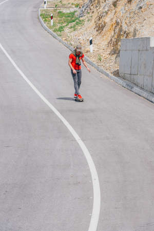 Male caucasian longboarder riding downhill on an empty road, preparing for a speed tuck on the next turn he has to make. Wearing a red t-shirt green hat and black jeans.の写真素材