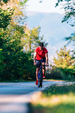 Fit cyclist rides his bicycle (bike) on an empty road in nature wearing a baseball hat and red t-shirt.の写真素材