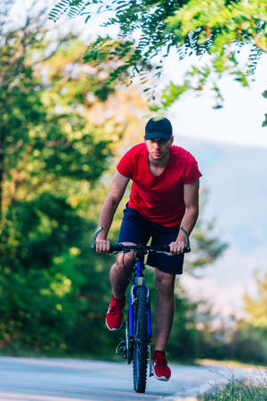 Fit cyclist rides his bicycle (bike) on an empty road in nature wearing a baseball hat and red...