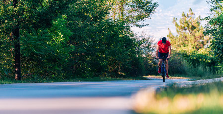 Fit cyclist rides his bicycle (bike) on an empty road in nature wearing a baseball hat and red...