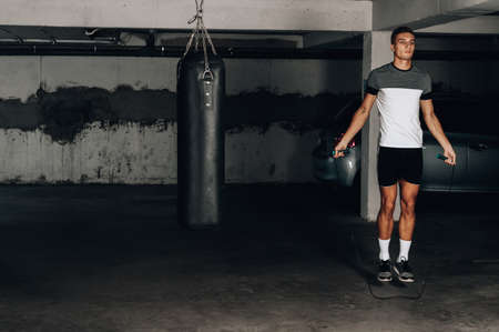 Masculine man skipping rope in dark. Portrait of muscular young man exercising with jumping rope