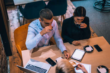 Top view of a beautiful young couple talking to their realtor while using tablet and drinking coffee at table in cafeの写真素材