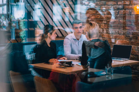 Business meeting inside a cafe with young colleagues sitting at a table, sharing some new ideas while the focus is through a cafe window.の写真素材
