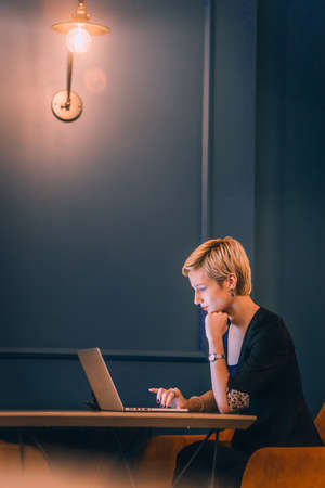 Confident young businesswoman working on her laptop while sitting in a corner of a cafe with dark blue wallsの写真素材