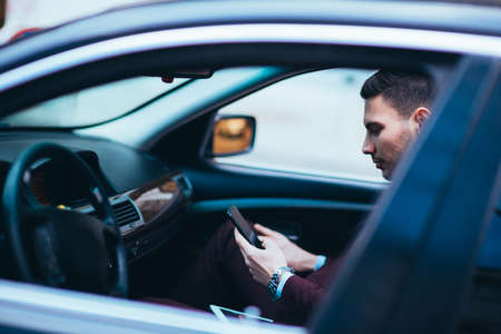 Young businessman looking at his cellphone while comfortably sitting at the front seat in his luxury...