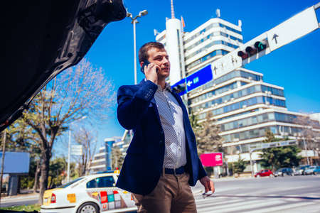 Young businessman is talking on his phone while looking under the lifted hood of his car.の写真素材