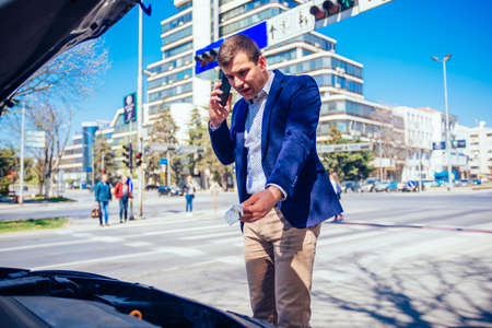 Young businessman is talking on his phone while looking under the lifted hood of his car.の写真素材