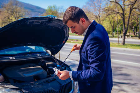 A handsome businessman wearing blue blazer lifting up the hood of his car and checking the oil level on a sunny day parked on a busy city boulevard.の写真素材