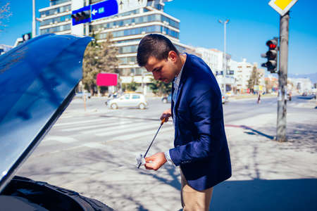 A handsome businessman wearing blue blazer lifting up the hood of his car and checking the oil level on a sunny day parked on a busy city boulevard.の写真素材