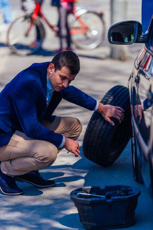 A formally dressed gentleman wearing a blue blazer is changing the flat tire on his car while being parked next to a busy boulevard.の写真素材