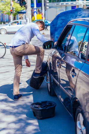 A sweaty tired young entrepreneur is wiping the sweat off his forehead while trying to change a flat...