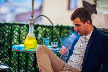 A handsome entrepreneur with facial hair smoking shisha(nargile) on his balcony while drinking...