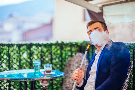 Young businessman wearing blazer and khaki smoking hookah at a coffee shop, while enjoying the amazing city landscape.の写真素材