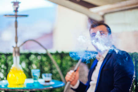 A handsome entrepreneur with facial hair smoking shisha(nargile) on his balcony while drinking coffee and enjoying the amazing view.の写真素材