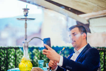 A happy businessman is smoking shisha (hookah) and looking at his phone while smiling and enjoying the beautiful sunny weather.の写真素材