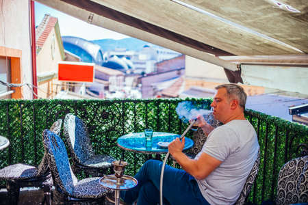 A middle-aged man dressed casually is smoking hookah (shisha) while sitting at an old wooden balcony...