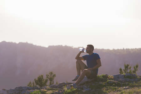 Handsome athlete sitting at a rocky peak while looking at the breathtaking mountain line and a beautiful lake while wearing a blue shirt and grey shorts.の写真素材