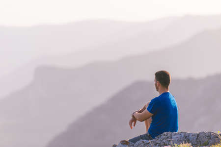 Adventurous man sitting on top of a mountain and enjoying the beautiful view, while looking downhill at the blue river and amazing mountain line.の写真素材
