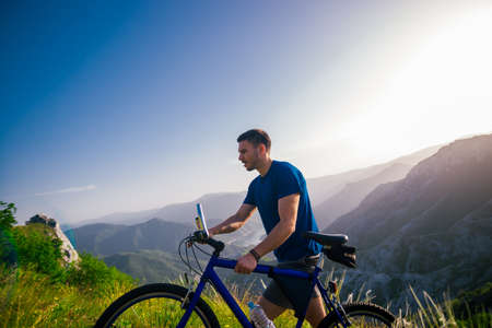 Perspective of a fit mountain biker pushing his bike uphill with amazing view on a forest, river and...
