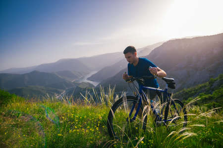 Perspective of a fit mountain biker pushing his bike uphill with amazing view on a forest, river and mountains in the background. Amazing green nature at sunset.の写真素材