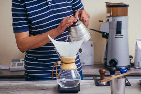 Close up image of young male barista pouring boiling water from kettle to drip coffee maker on...
