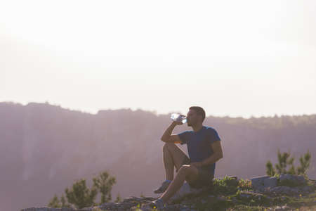 Handsome athlete sitting at a rocky peak while looking at the breathtaking mountain line and a beautiful lake while wearing a blue shirt and grey shorts.の写真素材