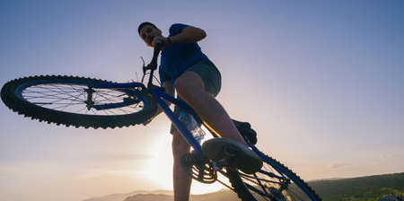 Strong fit male mountain biker performing stunts on rocky terrain on a sunset while wearing a blue shirt and riding a blue bikeの写真素材