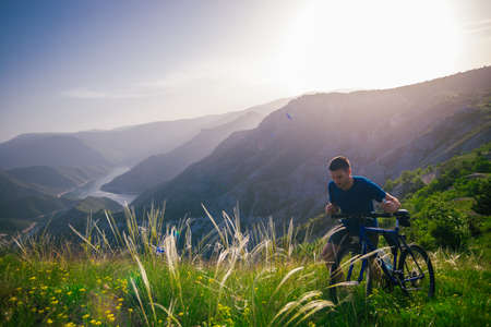 Perspective of a fit mountain biker pushing his bike uphill with amazing view on a forest, river and mountains in the background. Amazing green nature at sunset.の写真素材