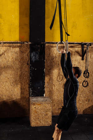 Portrait of young fit muscular man using gymnastic rings while exercising in gym