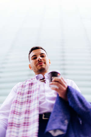 Handsome businessman is standing still, behind him is a big building, he is looking satisfied with a...