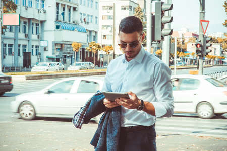 A stylish businessman is standing outside, looking serious with tablet in his hands