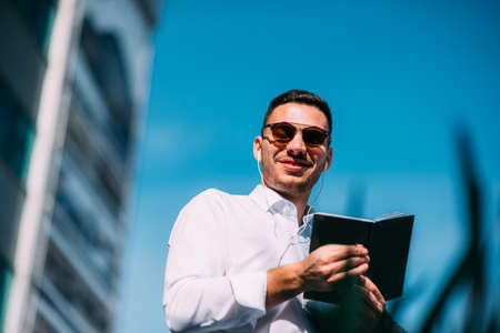 A businessman in suit is looking around him in the city while he holds a pen and opened notebookの写真素材