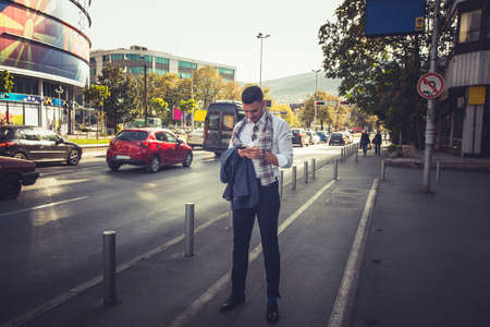 Happy businessman is standing on the sidewalk and looking at his phone, meanwhile he is enjoying the...